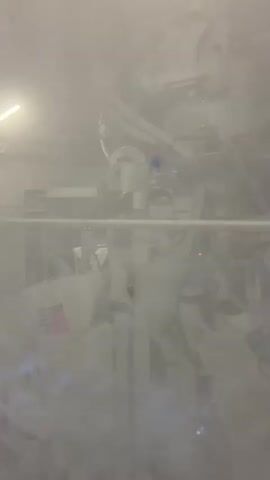The video shows a close-up of a white industrial machine, possibly a packaging or filling machine, in a brightly lit environment. The machine has various components including pipes, robotic arms, and conveyor belts. Several white bags with red and black lettering are visible, suggesting the machine is processing or filling these bags. The scene is slightly obscured by what appears to be mist or steam, creating a hazy atmosphere. There are no people visible in the frame. The camera appears to be static, capturing the continuous operation of the machinery.