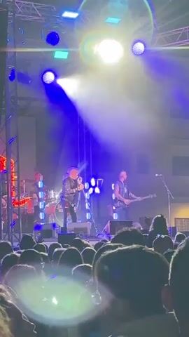 A band performs on a stage at night, with bright lights and smoke effects. The lead singer, a man with blonde hair, is in the center, singing into a microphone and wearing a black jacket. To his right, a man with dark hair plays a guitar. Behind them, a drummer is visible, as well as what appears to be a bass player to the far left. The stage is set up with lighting rigs, speakers, and large, illuminated letters that spell out "URT" on the backdrop. A crowd of people is gathered in front of the stage, many holding up their phones to record the performance. The overall atmosphere is energetic and lively.