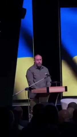 A medium shot captures a man at a wooden podium, addressing an audience. The man has a bald head and a graying beard, wearing an olive-green button-up shirt and what appears to be camouflage pants. His hands are clasped in front of him for a moment, then he gestures with them as he speaks into the microphone. Behind him, a large screen displays the colors of the Ukrainian flag, blue and yellow. The background is dark. The tops of heads of the audience are visible in the foreground. There is a white chair on the far right of the frame.