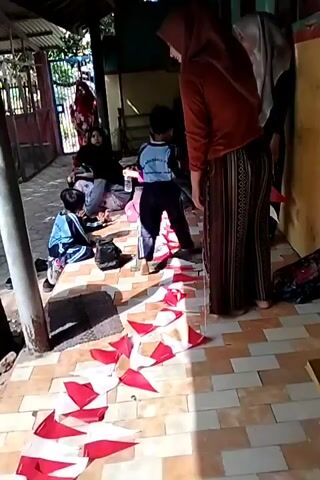 The video shows a group of children and adults gathered outdoors. Several children are seated on the ground, facing a row of small red and white flags laid out on the tiled floor. Two women, one wearing an orange hijab and the other a grey hijab, are standing and appear to be supervising or interacting with the children. One child, wearing a blue shirt and dark pants with blue stripes, is standing and looking towards the women. Another child, further down the row, is also seated. In the background, more people are visible, some sitting and some standing. The environment appears to be an open-air area with a roof structure supported by posts. A sink with a faucet is visible in the middle ground. The overall scene suggests a communal activity or event involving children and adults.