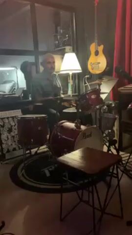 A man with a gray mustache and a patterned shirt sits behind a burgundy Pearl drum kit. He is in a room with a lamp, a keyboard, and a guitar hanging on the wall. A red curtain is visible on the right. The man is playing the drums.