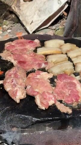 This is a close-up, overhead shot of raw pork being grilled on a dark, flat stone surface, possibly a griddle or a hot plate. Several pieces of thinly sliced pork, some with visible marbling and a pinkish-red hue, are arranged on the surface. To the right, there are several strips of what appears to be pork belly, with distinct layers of fat and meat, also raw and awaiting cooking. Some slight browning is beginning to form on the edges of the pork pieces, indicating they are starting to cook. Wisps of smoke are visible rising from the grilling surface, suggesting heat is being applied from below. In the background, out of focus, is a rustic, outdoor setting with what looks like dirt or gravel, a piece of white fabric or tarp, and some burnt material, possibly remnants of a fire. The overall impression is of a basic, outdoor grilling setup.