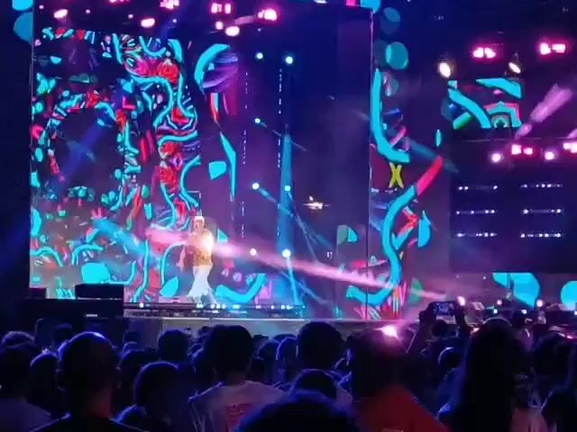 A concert stage is illuminated by vibrant, abstract digital graphics displayed on large screens behind a performer. The performer, wearing a light-colored hat, a patterned shirt, and white pants, stands on a raised platform and appears to be speaking or singing to a large audience. The audience is visible in the foreground, with many people holding up their phones, likely to record the event. The stage is also equipped with stage lighting, casting beams of light across the scene.