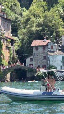 A boat with two people in bikinis is in the foreground, sailing on a body of water. In the background, a picturesque village is built into a hillside. Buildings with red-tiled roofs and ivy-covered walls are visible. A stone bridge arches over the water, and a crowd of people are gathered on top of it and on the banks. Some people appear to be swimming or wading in the water below the bridge. The scene is bathed in sunlight, suggesting a warm, summer day.
