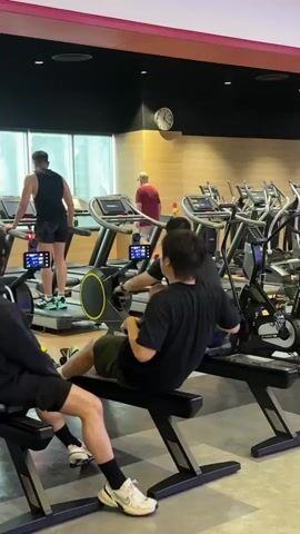 The video shows a gym interior with several people exercising. In the foreground, two individuals are seated on rowing machines, actively engaged in rowing. To the left and slightly behind them, another person is also on a rowing machine, partially visible. Further back and to the left, a man wearing a black tank top and shorts is walking on a treadmill. In the middle ground, a person in a red shirt is standing near a treadmill, and another person is seen walking on a treadmill to the right. The gym appears to be well-equipped with multiple treadmills and rowing machines. The lighting is bright, typical of a fitness facility, with warm-toned wood paneling on the walls and a black ceiling. A clock is visible on the wall in the upper right portion of the frame.