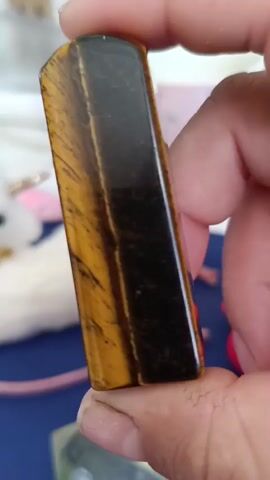 A close-up shot shows a person's hand holding a polished tiger's eye stone. The stone is rectangular with rounded edges and features bands of golden yellow and dark brown. The hand is holding the stone at an angle, allowing light to reflect off its smooth surface. In the background, out of focus, there are hints of other objects, including what appears to be a white fluffy item and a pink string. The lighting suggests an indoor environment.