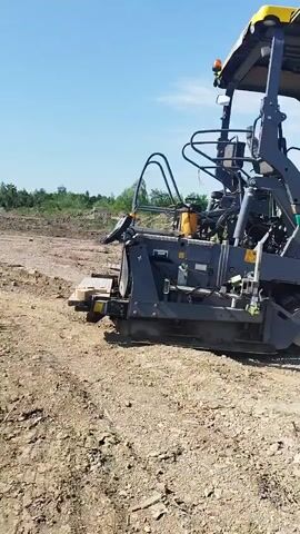 This media file shows a close-up, side-view of a green and black Vogele Super 1900-3 asphalt paver on a construction site. The machine is stationary, with its tracks resting on dry, dusty ground. In the background, there is a clear blue sky with some scattered clouds, suggesting it is daytime with fair weather. The surrounding environment appears to be undeveloped land, with some scrubby vegetation and a partially constructed building visible in the far right background.

The paver is a large piece of construction equipment, characterized by its yellow canopy, the operator's seat, and the extended screed at the front where asphalt would be laid. The text "SUPER 1900-3" is clearly visible on the side of the machine. The overall impression is of a construction project in progress under good weather conditions. There are no people visible in the frame. The scene conveys a sense of industrial activity and readiness for work. The location is identified as Rozdil, Ukraine, which provides geographical context to the construction setting.