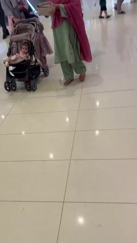 A person is pushing a stroller with a baby in it through a mall. The stroller is black and silver. The baby is wearing a pink and white outfit. The person pushing the stroller is wearing a pink shawl and a green and white outfit. The mall has tiled floors and escalators. There are other people in the mall, including a child walking with an adult and people standing near an escalator.