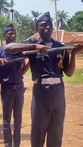 The video captures a group of young men in blue uniforms performing a military-style drill outdoors under bright daylight. Each individual wears a short-sleeved blue shirt, dark trousers, and a dark garrison cap. They are equipped with wooden prop rifles, which they hold and maneuver during the drill.

The main subject in the foreground, whose name tag reads "MC LUKAS," stands prominently with a white belt and has multiple medals or ribbons pinned to his uniform. The drill begins with a clear command, "Shoulder arms! Up!" followed by the participants smoothly transitioning their wooden rifles from a horizontal position to a vertical "shoulder arms" stance. Their expressions are focused and serious, indicating concentration on the drill.

The setting features a dirt ground, with clear skies and strong sunlight creating distinct shadows. In the background, palm trees and other lush foliage are visible, alongside distant buildings, suggesting a rural or semi-rural environment. Briefly, behind the uniformed line, a young person in a white dress and green top, wearing a crown, can be seen. The scene is consistent with an outdoor event in a tropical or sub-tropical region, such as Andaha, Nigeria.