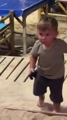A toddler stands in sand. He wears a gray shirt and dark blue shorts. He holds a black object in his left hand. Behind him are tables and chairs, some covered with blue tablecloths. A wooden walkway is partially visible.