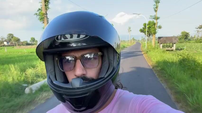 A man wearing a black motorcycle helmet and pink shirt is riding a motorcycle on a paved road. The road is lined with green grass and trees. In the background, there are houses and fields. The sun is shining brightly, casting shadows on the road. The man is looking at the camera, and his face is partially visible through the helmet. He appears to be smiling. The video is shot from the perspective of the man, giving the viewer a first-person view of his journey.