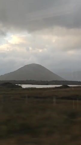 The video shows a landscape view from a moving vehicle. A large, rounded mountain dominates the background under a cloudy sky. In the middle ground, a body of water stretches across the scene, bordered by dark green and brown vegetation. The foreground consists of blurred green and brown foliage, with a yellow barrier visible at the bottom. The overall atmosphere is overcast and muted.