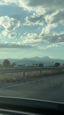 A car's interior dashboard is visible in the foreground. The video captures a highway scene from the perspective of a moving vehicle. The road is paved and has multiple lanes. A metal guardrail separates the highway from the grassy shoulder. In the distance, a range of mountains with snow-capped peaks is visible under a bright sky with scattered clouds. Several cars and trucks are on the highway. Some trees and structures are present along the roadside. The lighting suggests it is daytime, possibly late afternoon or early morning.