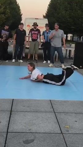 A group of people watch a breakdancer perform on a light blue mat. The dancer, wearing a white cropped shirt and black pants, is in motion, with their left arm extended and their body twisted. Several men stand behind the dancer, some with microphones and one wearing a baseball cap and a jersey with the number 32. A white van with an orange logo is parked in the background, and trees line the area. The ground around the mat is paved with grey stones.