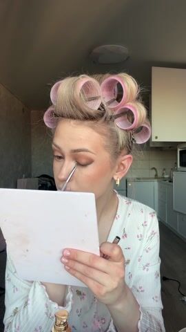 A woman with blonde hair in rollers applies makeup in a brightly lit apartment. She wears a white floral-patterned robe and gold earrings. She holds a white compact mirror in her left hand and uses a brush in her right to apply eyeshadow to her left eye. After applying the eyeshadow, she looks at the camera and smiles slightly. The apartment includes a kitchenette visible in the background.