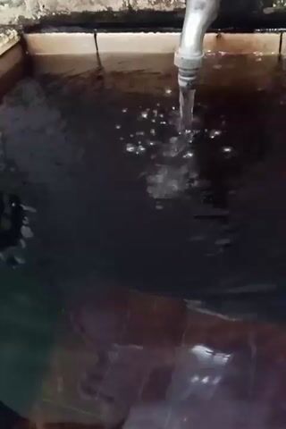 A faucet is pouring water into a dark, square container, creating ripples and bubbles on the surface. The container appears to be made of wood or plastic. The water is dark, and the bottom of the container is visible through the water, appearing to be tiled or covered in a patterned material. The camera is angled downwards, focusing on the water and the faucet.