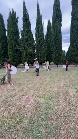 A group of women in dresses are gathered in a grassy area with tall cypress trees in the background. Some of the women are holding large white paper umbrellas or fans. One woman in the center appears to be directing the others. The sky is overcast. The overall impression is of a staged photo shoot or a celebratory event.