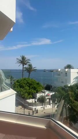 The video shows a view from a balcony or window overlooking a tropical coastal scene. The foreground features a glass railing and part of a white building. Beyond that, there are several palm trees, lush green foliage, and a body of blue water. In the distance, several rocky islands or outcroppings are visible in the sea. There are also white buildings with balconies along the coastline. The sky is a clear blue with a few wispy clouds. The overall impression is a bright, sunny day in a seaside resort.