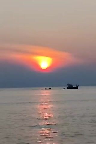 The video shows a sunset over the ocean, with the sun appearing as a bright yellow-orange orb partially obscured by clouds. The sky is a gradient of colors, transitioning from greyish-blue near the horizon to a mix of orange, pink, and grey higher up. The sea is relatively calm with faint ripples and a subtle shimmer reflecting the colors of the sunset. Two boats are visible in the distance on the water. One boat appears smaller, further away, and is silhouetted. The other boat is larger and closer, with a visible cabin and details suggesting it's a fishing vessel. The overall scene is serene and depicts a natural landscape at twilight.