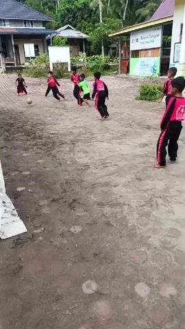 A group of barefoot children wearing matching athletic gear consisting of black bottoms and pink and black tops play soccer in a muddy outdoor setting. The children are near a white tent and are playing on a patch of dirt with a few patches of brick flooring. In the background is a two story building with a gabled roof and other smaller buildings and trees. One child wears a green shirt.