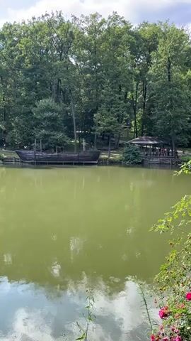 A video shows a calm, green lake. On the far bank, a wooden ship, appearing to be a replica, is moored near a wooden dock and a covered pavilion. Trees line the background, and foliage is visible in the foreground. The sky above is partly cloudy.
