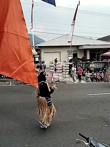 The video shows an outdoor street scene, likely a parade or cultural event. Several people are dressed in traditional, grass-like skirts and headpieces, performing a dance. Some are also wearing dark tops and white shoes. In the background, there are buildings, market stalls, spectators, and decorative flags. An orange banner is visible on the left side of the screen. The ground is asphalt, suggesting a road. The sky is overcast. A motorcycle is parked in the foreground, partially obscuring the lower part of the frame.