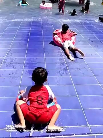 The video shows a group of children enjoying themselves in a shallow outdoor pool on a sunny day. The pool has a blue tile floor and gentle water jets create small ripples on the surface. Most of the children are wading or sitting in the water, some using inflatable rings for support. One child in a red swimsuit is attempting to do the splits on the tiled edge of the pool, with an inflatable ring around their waist. The other children seem to be playing and splashing around, with a few adults visible in the background, presumably supervising. The atmosphere appears lighthearted and playful. The bottom of the screen displays information from the device used to record the video: "OPPO A6 Pro fataklumy 30 Mei 2023 pukul 11:28 Kabupaten Bireuen."