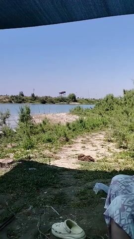 A woman with dark hair, wearing a light blue tank top and a patterned skirt, sits in a chair under a canopy, smoking a cigarette. She is facing towards the right of the frame. In the background, there is a body of water with several fishing rods set up along its edge. The area is covered with dry grass and bushes, and the sky is clear blue. A plastic container with various items is visible on the ground to the right of the woman.