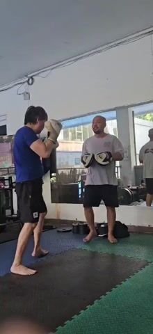 The video shows two men in a gym. The man on the left is wearing a blue t-shirt and black shorts, and has boxing gloves on. He is punching towards the man on the right. The man on the right is wearing a grey t-shirt and black shorts, and is holding boxing mitts. He is looking at the man on the left and appears to be coaching him. In the background, there is a mirror reflecting other people and a swimming pool. The gym has padded flooring and weights are visible.