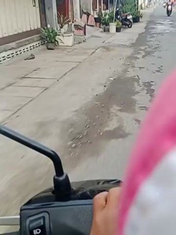 The video shows a first-person perspective from a motorbike traveling down a paved road. On the left side of the road are buildings with shops, some with potted plants and motorcycles parked outside. There are also Indonesian flags visible, suggesting a national celebration. The road itself appears wet in patches, indicating recent rain. Several other motorbikes are seen in the distance, also traveling down the road. The sky is overcast.