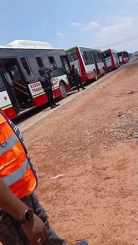 A group of white and red buses are parked in a line on a dirt road. A person in an orange vest is standing in the foreground. Some of the buses have their doors open and people are standing near them. The sky is blue with some clouds. The ground is covered with dirt and gravel. The buses appear to be part of a public transportation system.