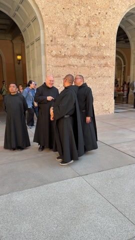 The video shows a group of four men in black robes standing in an arched hallway. They appear to be monks, and they are in conversation. One man is smiling and pointing. The camera pans up to show a building with a rough, stone facade with arched openings and a small window, an outdoor light fixture, and carvings above. Several people are visible in the background, walking through the archways.