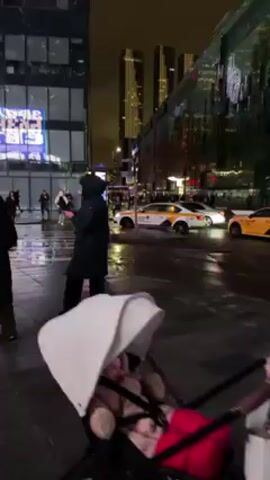 A woman pushes a baby stroller across a wet city street at night. Another woman stands nearby taking photos with her phone. A man walks by. There are several yellow and white taxis on the road. Tall buildings with bright lights line the street. Pedestrians can be seen walking on the sidewalk.