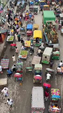The video shows a busy street market scene. Numerous people are present, walking and interacting within the crowded space. Various modes of transportation are visible, including numerous rickshaws, some of which are carrying goods, and a few motorcycles. The market stalls are laden with fresh produce, primarily fruits like mangoes and what appear to be dragon fruits, displayed on carts and on the ground. Some of the rickshaws and stalls are covered with colorful tarpaulins. The environment appears to be urban, with buildings visible on either side of the street, and a bridge overhead. The overall impression is of a bustling and vibrant marketplace.