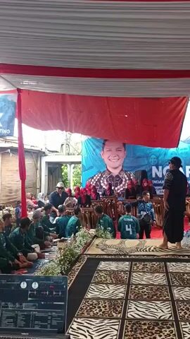 The video shows a group of people gathered under a large tent, possibly for a community event. There are men and women, some sitting on the floor, others standing and walking around. Many are wearing green shirts, suggesting a uniform or coordinated outfit. A laptop with DJ software is visible in the foreground, indicating music or audio is being played. Banners are hung behind the crowd, featuring images of people and text, one visible banner reads "NGAPRAK" and "Mendak Keluarga Berisiko Stunting". The setting appears to be semi-outdoors with buildings visible behind the tent.