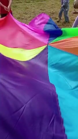 The video shows a group of children and adults gathered outdoors on a grassy area. In the foreground, a large, colorful parachute, segmented into sections of purple, teal, green, yellow, pink, and orange, is being manipulated. Several children are actively holding onto the edges of the parachute, pulling and lifting it. In the background, more people are visible, some standing and others seated at tables. A red tent or canopy is set up in the middle ground, and trees and a building are in the far background. The overall scene appears to be a casual outdoor gathering or event.