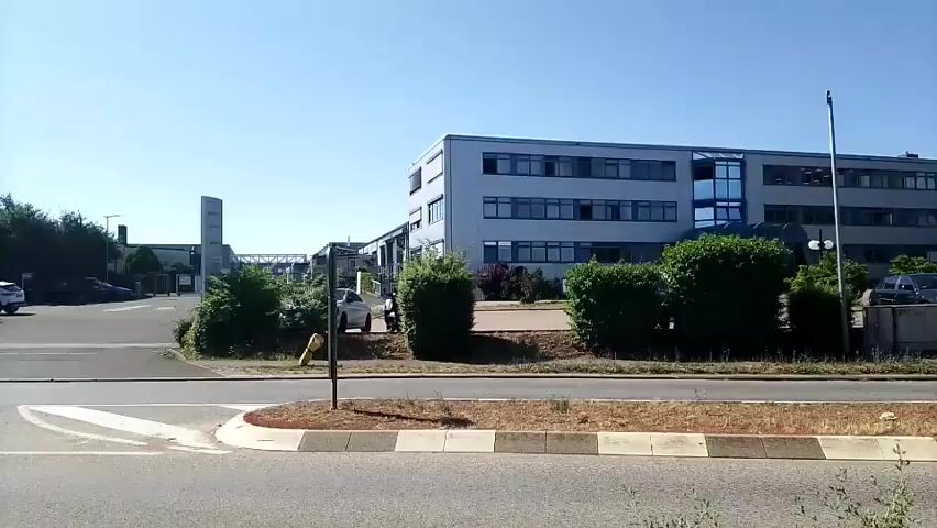 The video shows a street with a building on the right and trees and other buildings in the background. The sky is clear and blue. In the foreground, there is a road with a curved sidewalk and a small garden area. A few cars are visible on the road and in the parking lot of the building on the right. One car is driving past the camera on the left side of the frame. The building on the right is a multi-story office building with a modern design, featuring large windows and a gray facade. There are some bushes and trees around the building. The overall impression is of a typical suburban or business park setting on a sunny day.