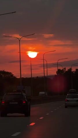 The video shows a view from a car driving on a highway at sunset. The sun is a bright orange ball partially obscured by clouds, casting a warm glow across the sky. Several cars are visible on the road, all moving in the same direction. Streetlights line the highway, and trees can be seen in the background. The overall impression is one of a peaceful, scenic drive as the day ends.