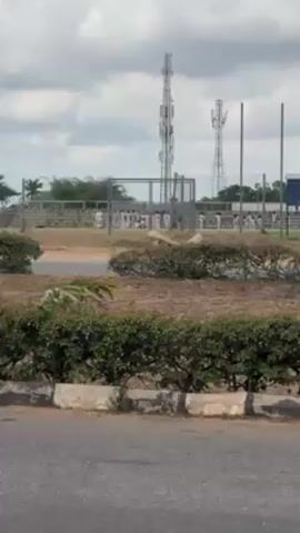 The scene shows a sports field from across a road lined with greenery. A group of people, mostly dressed in white, are visible on the field, performing what appears to be synchronized marching or drills. There are goalposts visible on the field. Behind the field are stadium bleachers. In the background, two tall telecommunications towers stand against a cloudy sky. The camera is static.