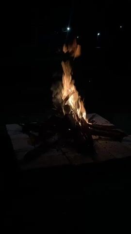 A fire is burning outdoors at night. The fire is contained on a stone slab and is made of wood. The flames are tall, and the area surrounding the fire is dark. The light from the fire illuminates the slab. There are lights in the distant background. There are faint voices heard.