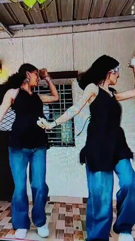 Two young women are dancing together outdoors. They are wearing matching black tank tops and blue baggy pants. The woman on the left is wearing glasses. They are holding hands and moving in sync, with their arms raised and bent. They are standing on a tiled patio. The background consists of a wall with a window and some hanging plants. The lighting suggests it is daytime.