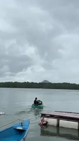 A motorboat with three occupants is traveling away from the camera on a body of water. The boat is green and the people are mostly obscured from view. The water is calm with a slight wake behind the boat. A mangrove forest lines the distant shore, and a small mountain or hill is visible in the background. In the foreground, a blue boat is docked to the left, and a wooden dock with white pylons is on the right. The sky is overcast with dark clouds.