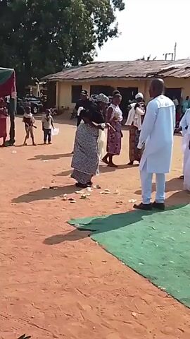 Outside on a sandy ground, a group of people are gathered. Several are dressed in white traditional attire. One woman in a patterned skirt and dark top dances in the center, throwing what appears to be money. Other people watch and some film with their phones. A tent is visible on the left and a low building is in the background. The sky is bright, and there are trees to the left.