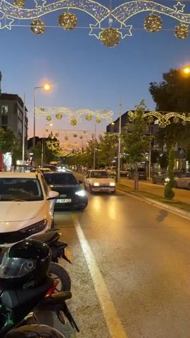 The video captures a street scene in Osmaniye, Turkey, during twilight. The sky is a deep blue, indicating dusk or early evening. Decorative yellow string lights, shaped like ribbons and stars, are strung across the street, suggesting a festive atmosphere, possibly related to a holiday or special event.

The street is lined with buildings, some of which appear to be shops or restaurants, indicated by glowing red and white signage. Trees with green foliage are planted along the sidewalk. Several vehicles are present on the road, including cars, a white van, and motorcycles. A black car is in the foreground on the left, partially obscured. A white car is driving down the street, and another white car with a license plate that reads "46 KK 203" is seen from the side. Further down the road, a white car's taillights create streaks of light, suggesting motion.

The activity appears to be normal street traffic and pedestrian activity, set against the backdrop of evening lights. The overall mood is calm and visually appealing due to the contrast between the darkening sky, the warm glow of the streetlights and building lights, and the decorative illumination. No people are clearly discernible due to the distance and motion blur. The presence of decorative lights suggests a celebratory or enhanced visual setting for the urban environment.