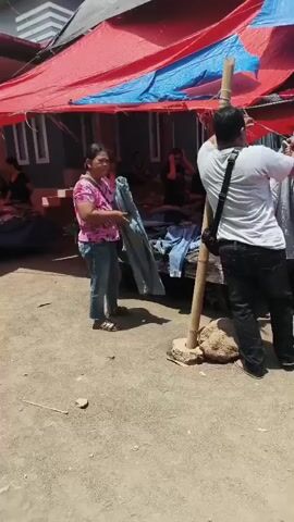 The video shows an outdoor market scene. Several people are present, mostly gathered under a red and blue tarp for shade. In the foreground, a woman in a pink floral shirt and jeans is holding up a piece of light blue fabric, possibly clothing, and looking towards a man who is partially visible on the right side of the frame. He is wearing a white shirt and dark pants, with a black bag slung over his shoulder. He appears to be interacting with a large bamboo pole. Further back, a young person in a red hoodie and shorts is holding a bunch of green vegetables in a black plastic bag. Other individuals are visible in the background, some behind tables or blankets displaying goods, suggesting a marketplace setting. The ground is dirt and gravel. The overall atmosphere appears to be casual and active, typical of a local market.