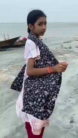 A young woman is seen from behind, walking along a grey, rocky beach toward the ocean. She is wearing a light pink top, maroon pants, and a dark, floral print shawl draped over her shoulders. The shawl flows behind her as she walks. The sky is overcast, and the water is a dull grey. In the background, there are small boats and other people on the beach.