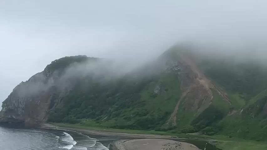 The video shows a drone shot of a foggy, mountainous coastal landscape. The drone flies forward, revealing green, forested hillsides partially obscured by mist and fog. In the foreground, waves crash on a sandy beach. There are no people or animals visible.