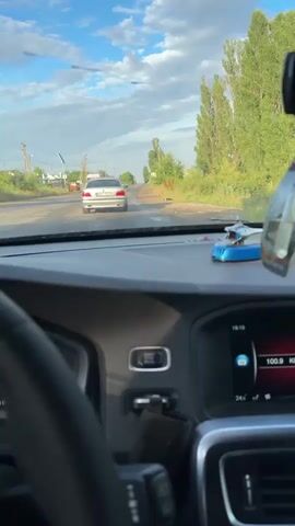 The video is filmed from the perspective of a driver inside a car. The driver's steering wheel and part of the dashboard are visible in the foreground. On the dashboard, there is a small toy helicopter and a screen displaying the time and temperature. The car is on a paved road, with other vehicles ahead. The road is lined with trees and vegetation on the right side. The sky is blue with some clouds, and the sun appears to be low, suggesting it might be late afternoon or early evening. There are some white streaks on the windshield, possibly from dirt or rain. The overall environment appears to be rural or suburban.