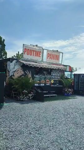 The video pans across an outdoor fair or festival. In the foreground, a gravel-covered area is visible. To the left, a food truck is parked, advertising "POUTINE" and "PANINI" with a cartoon chef illustration on its side. A menu board displays various food items. Beside the food truck, potted plants add decoration. Further to the right, a white tent with red accents is set up, possibly a vendor booth or seating area, with a red Coca-Cola branded refrigerator visible. Beyond the tent, a Ferris wheel is prominent in the background, indicating an amusement park or fair setting. A large yellow tractor is parked to the right of the Ferris wheel, with a large white "EXPO" sign behind it. Several people are walking around in the background, some near the tractor and others further away. The sky is blue with scattered clouds.