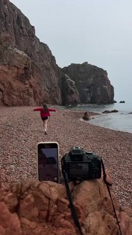 A young woman in a red jacket and black shorts walks toward the camera on a pebble beach, against a backdrop of tall, dark red cliffs and a gray sky. A smartphone and a DSLR camera are placed on a reddish-brown rock in the foreground. The smartphone screen displays a video recording of the scene. The ocean is visible to the right.